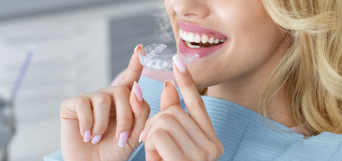 blonde woman with healthy white teeth holding invisible braces while sitting at dental chair, closeup portrait