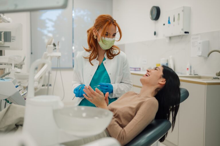Female dentist wearing surgical mask holding dental tools talking to happy female patient sitting in dentist chair in bright modern dental clinic
