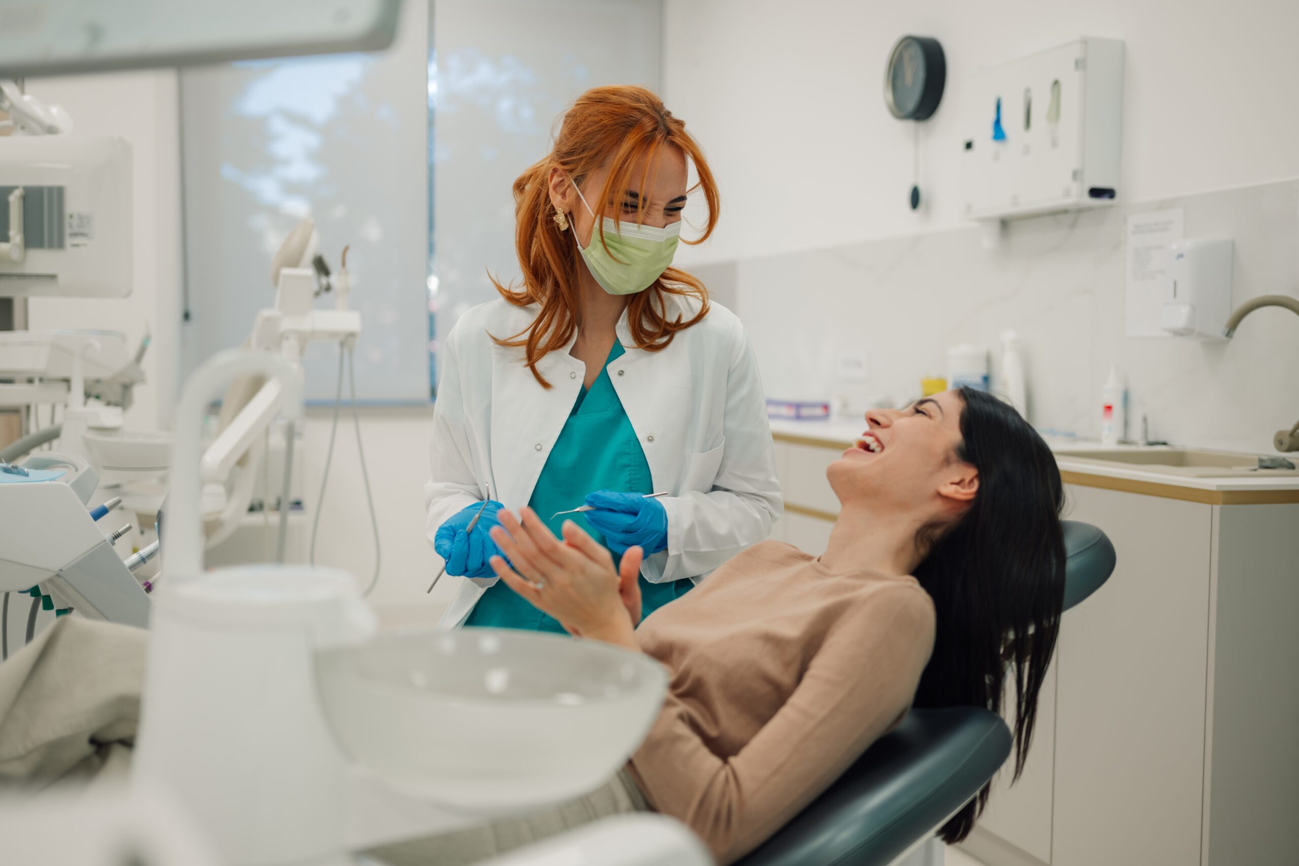 Female dentist wearing surgical mask holding dental tools talking to happy female patient sitting in dentist chair in bright modern dental clinic