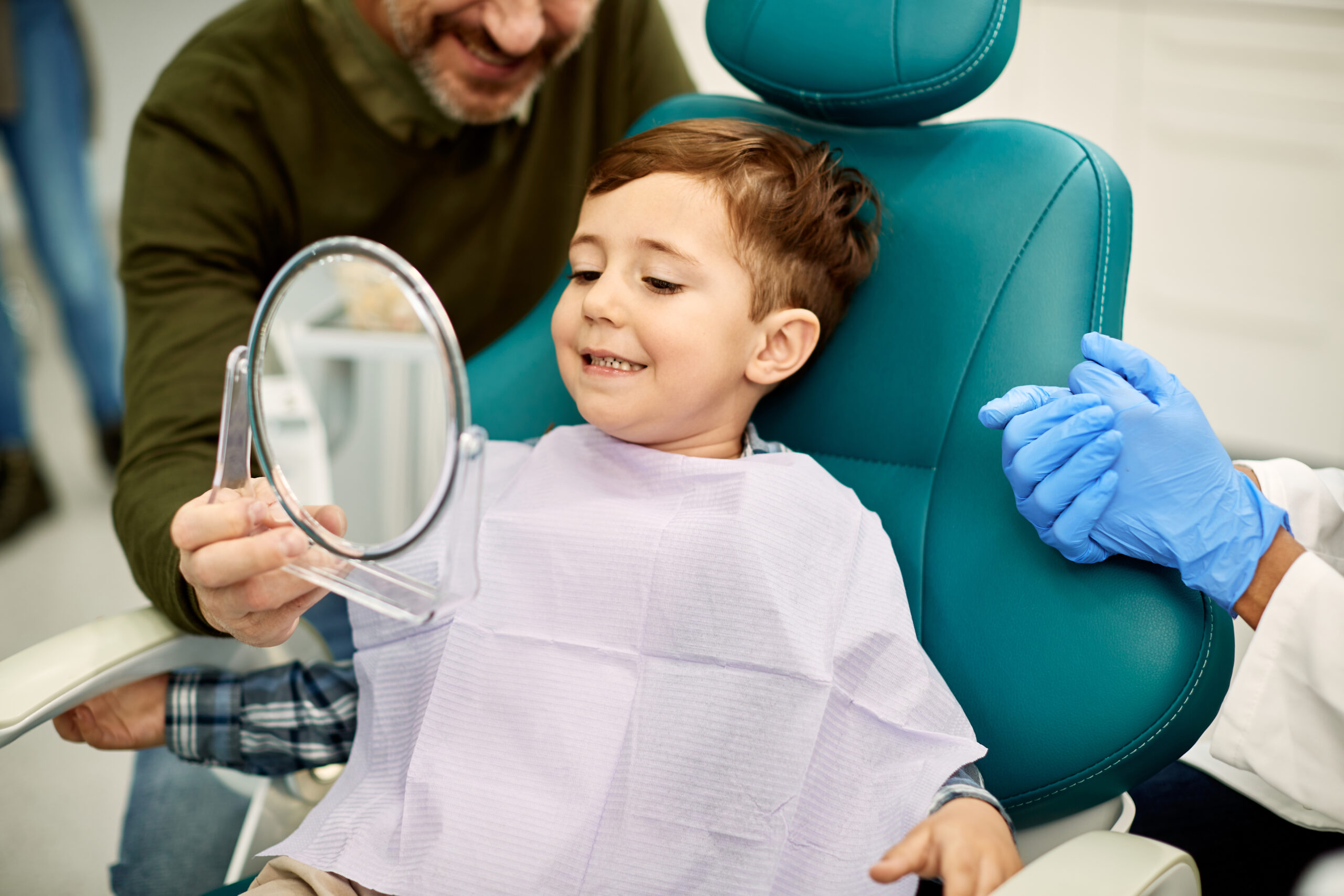 Smiling little boy holding a mirror and looking at his teeth